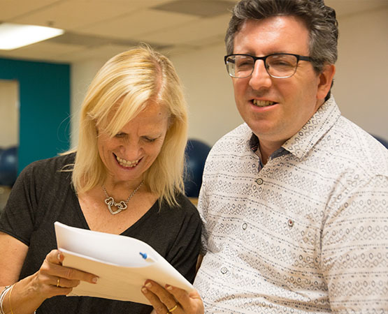 Blond womand in a black shirt smiling and looking at a script next to a smiling man in a white printed shirt
