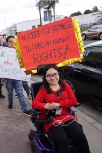 Mariela attending a rally for disability rights wearing a red long-sleeve shirt and holding a sign that says "#Keep the promise to fund my human rights!"