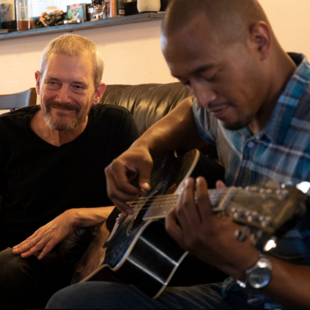 Rob smiling while listening to another man playing the guitar next to him