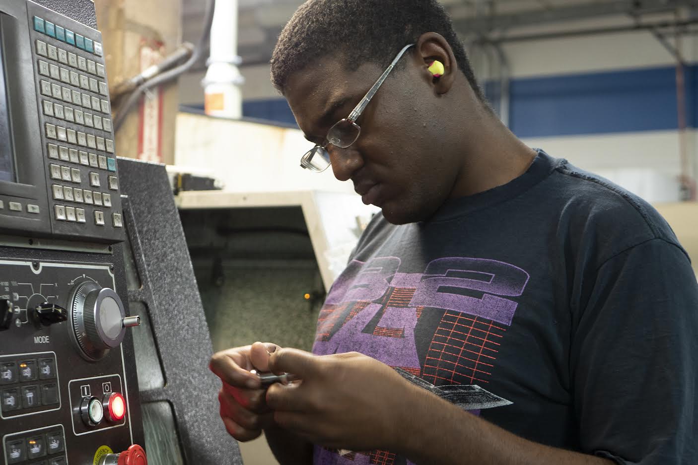 A young African American male student examining a piece of equipment in his hand while standing next to a control consol at a CNC machining shop during a Uniquely Abled Academy (UAA) training.