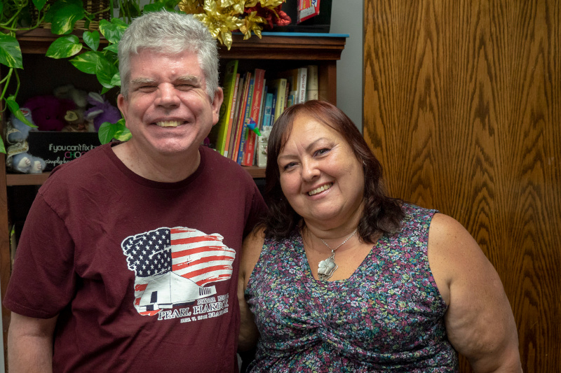 Older man with grey hair and a burgundy t-shirt and a woman with brunette hair and a floral blouse smiling while standing in front of a bookshelf in an office