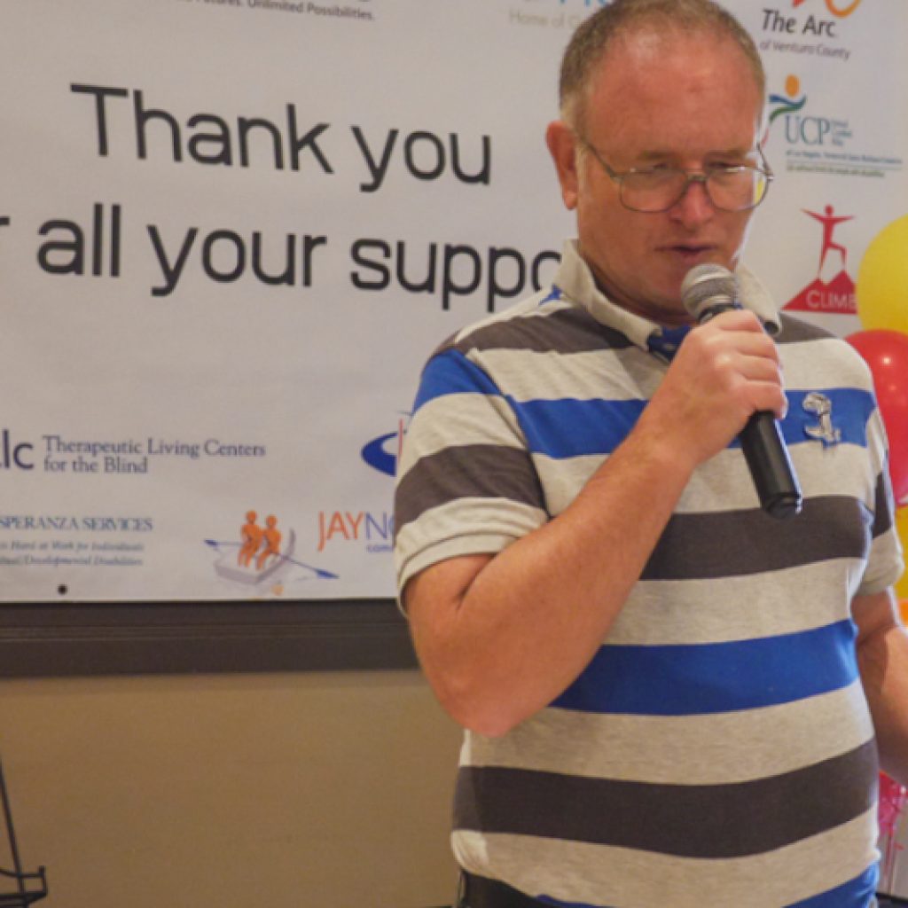 Man in a striped shirt giving a speech infront of a sign that reads "Thank You For Your Support"
