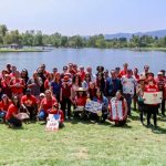 A group photo in front of a lake of around 40 people wearing red and holding signs supporting autism acceptance