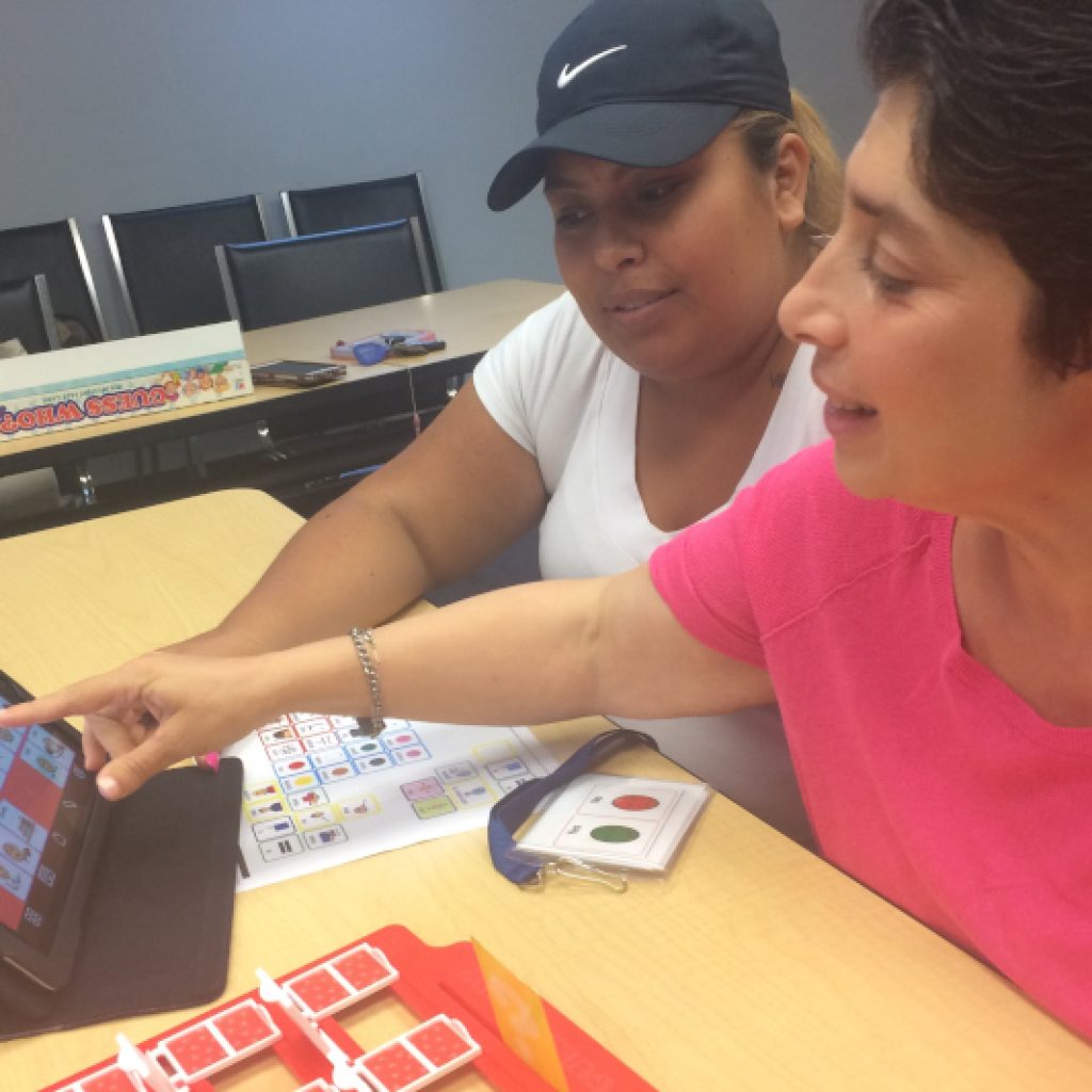 A woman in a pink shirt poining to a screen on an assissted communications device to show something to another woman wearing while sitting next to her.