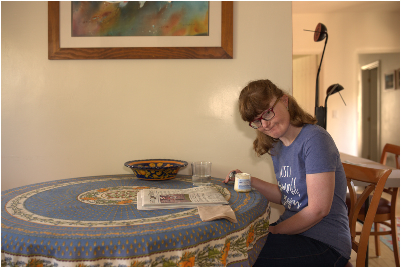 Adult woman supported through Jay Nolan SL services having a coffee and reading a newspaper at a table in her home.