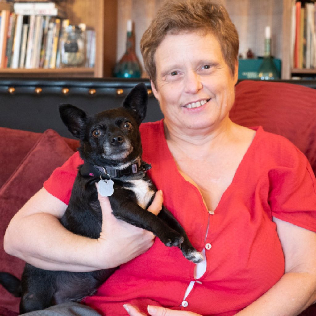 A woman in a red shirt sitting on a couach and holding a small black dog.