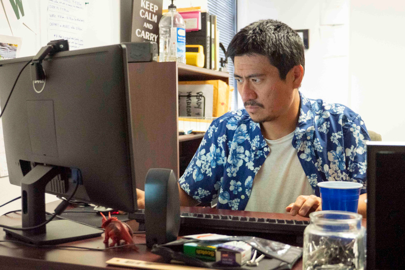 A young asian man in a hawaiian shirt working at the computer in his office.