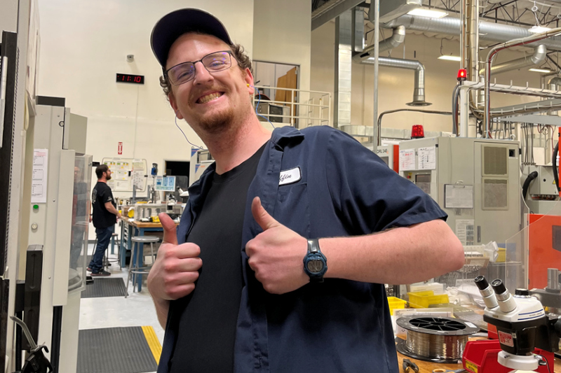 A graduate from UAA holding up two big thumbs up while on the job at a CNC machine workshop where he works.