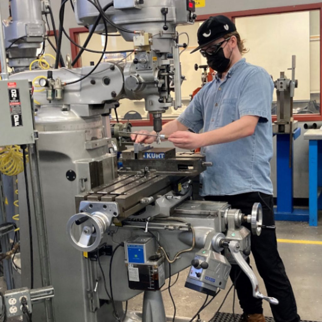 A young cucasian man in a jean button down shirt using a machine in a machining workshop