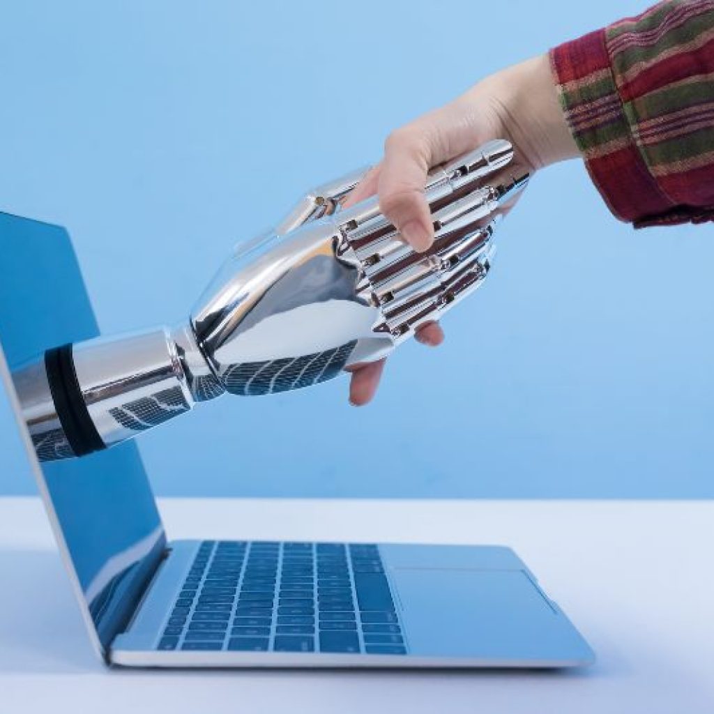 An artistic representation of AI: A robotic hand extending out of a laptop computer to shake hands with a human.