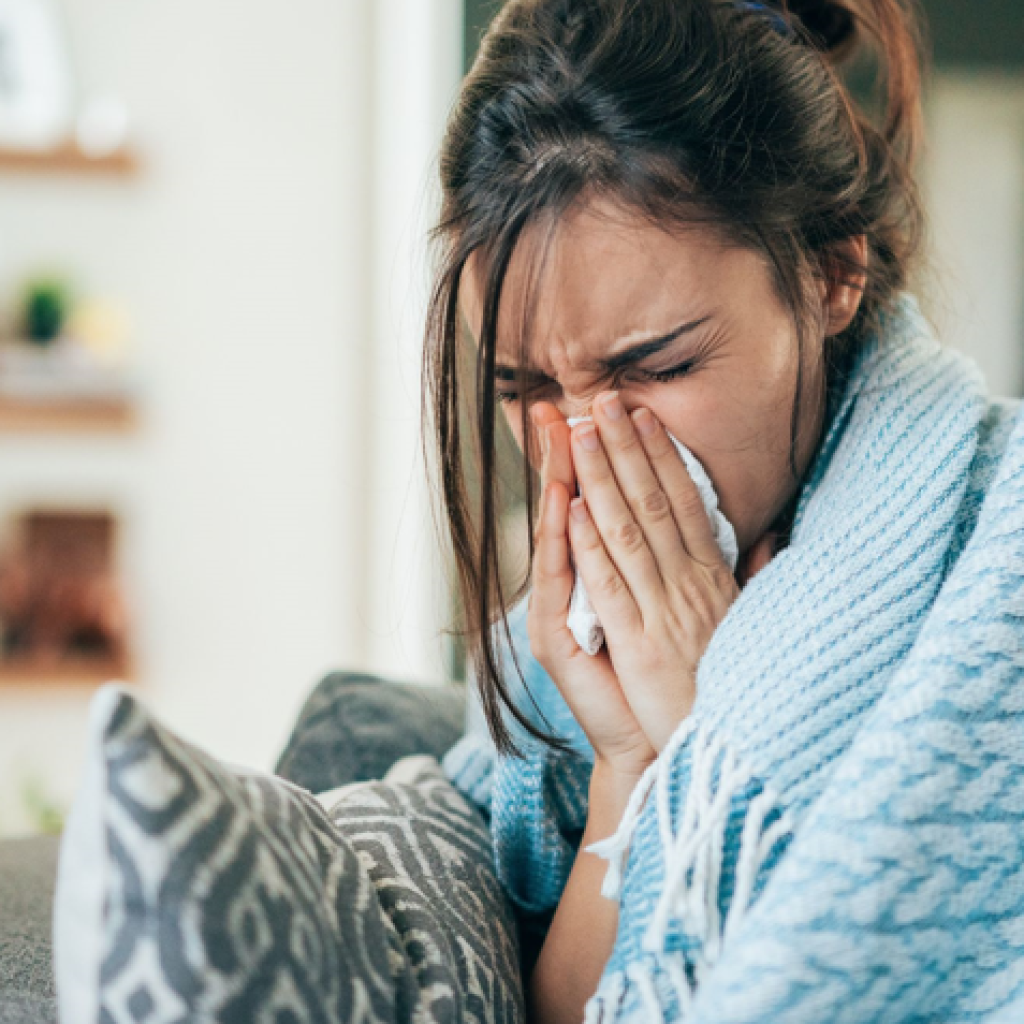 A woman wrapped in a blanket sneezing into a tissue, appearing to have a cold or allergies.