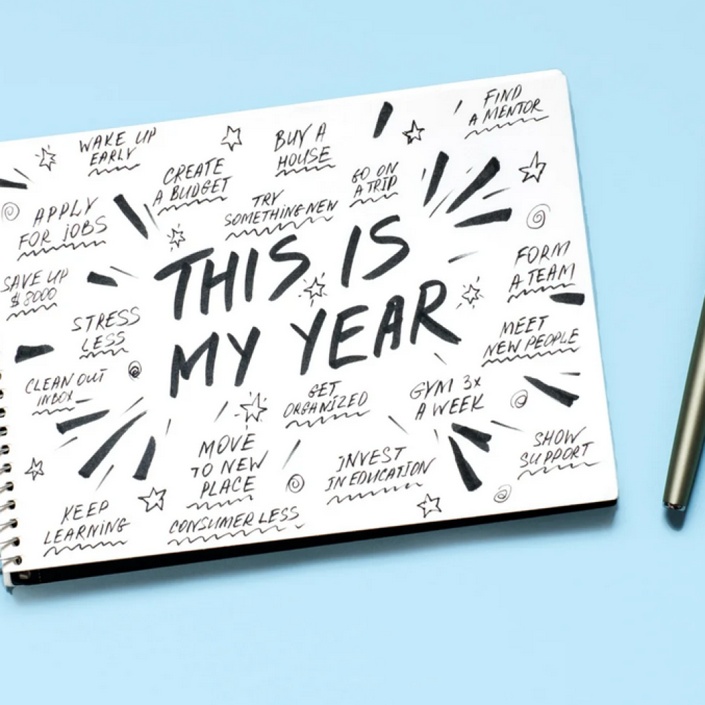 A notebook with "This is My Year" written in large letters, surrounded by various goal-related phrases, and a pen placed beside it on a light blue background.