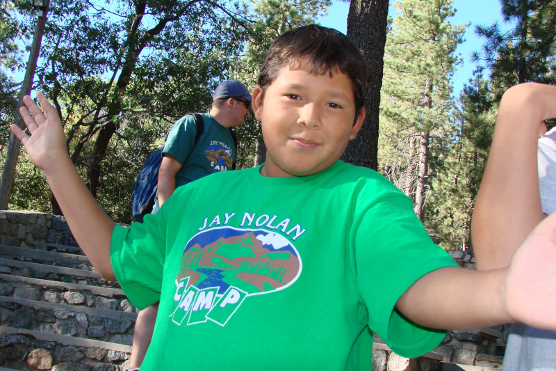 A young boy in a green Jay Nolan Camp T-shirt smiling.