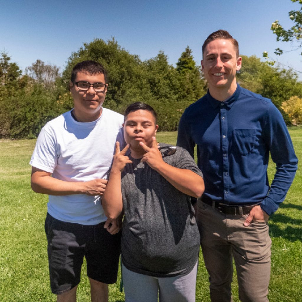 Three people standing together in a park, with one making a peace sign, and all smiling.