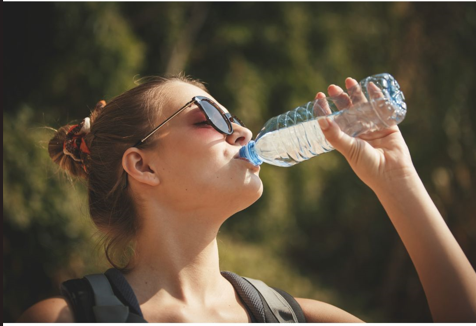 Mujer caucásica con gafas de sol bebiendo una botella de agua en un día caluroso.