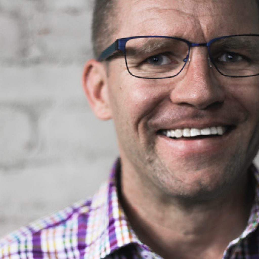 Portrait of JNCS Board Member Jason Freeman. He is a Caucasian middle-aged man wearing glasses and a purple checkered shirt.