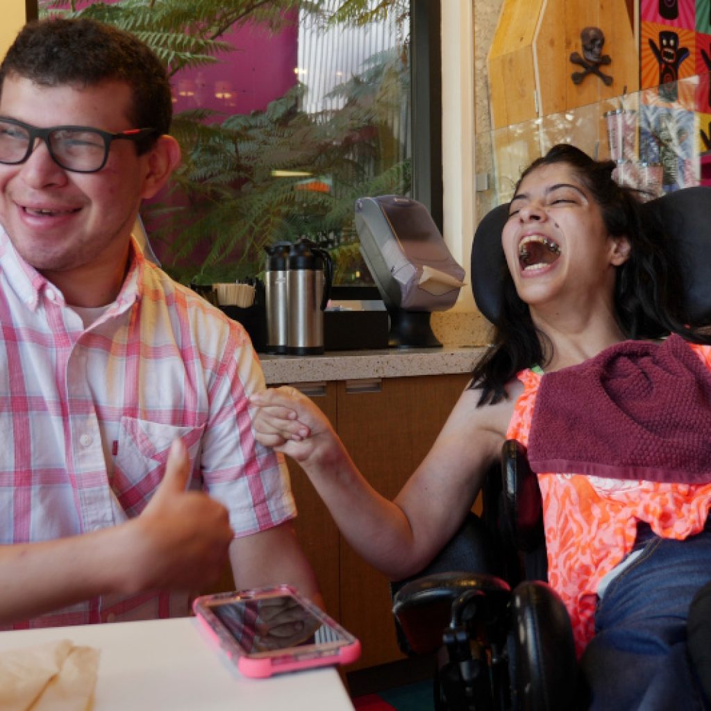 A Caucasian man wearing a pink checkered shirt is sitting at a table holding up a thumbs up sign. Next to him, his girlfriend is in a wheelchair and smiling widely.