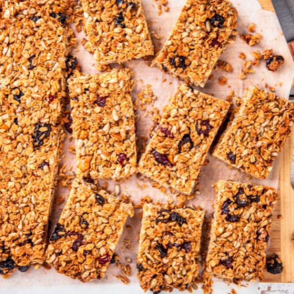 A batch of homemade granola bars cut into rectangles, placed on parchment paper on a wooden cutting board. A knife with a wooden handle is also on the board. The granola bars appear to be filled with nuts, seeds, and dried fruits.