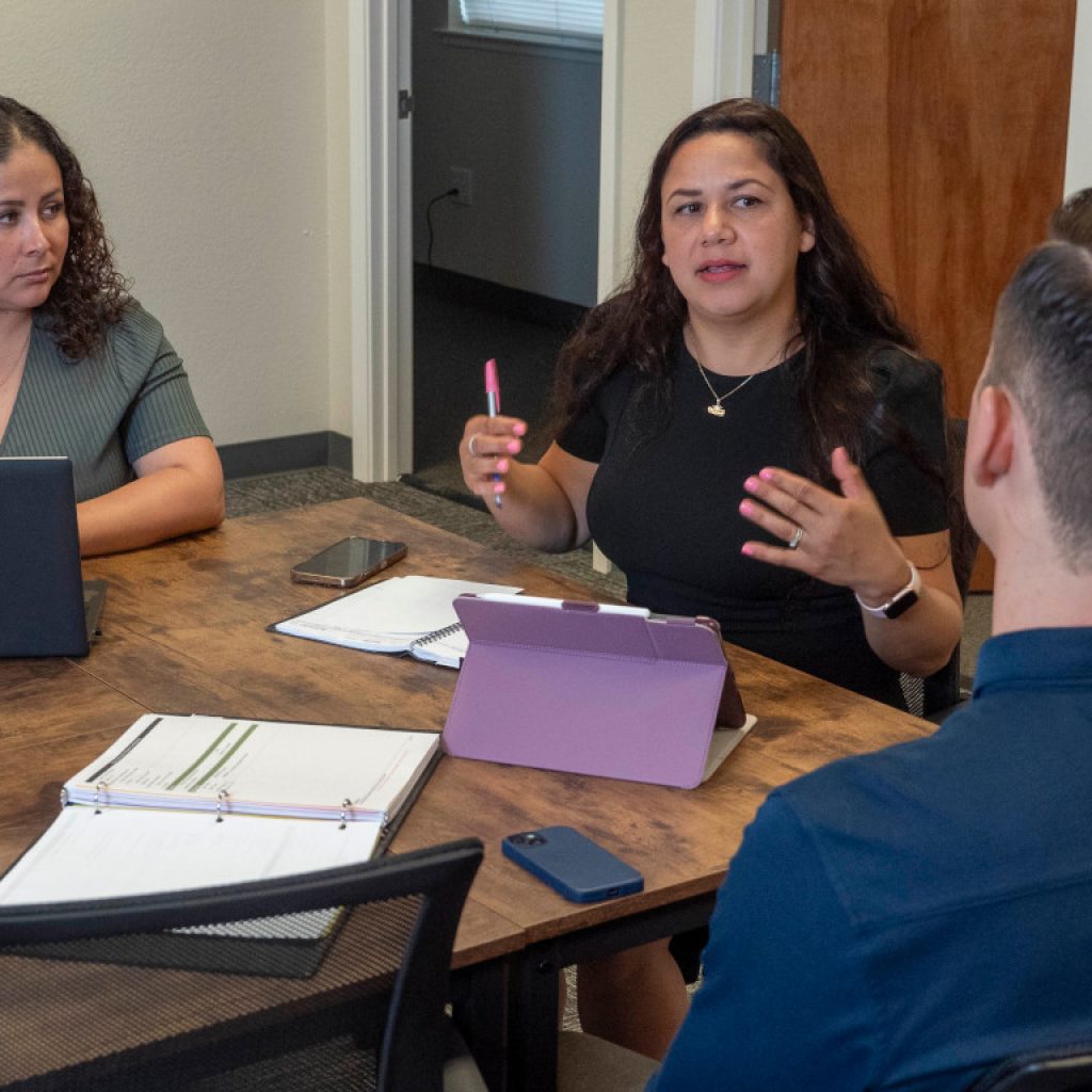 Three people sit around a table during a meeting, engaged in a discussion, with two of them using laptops.