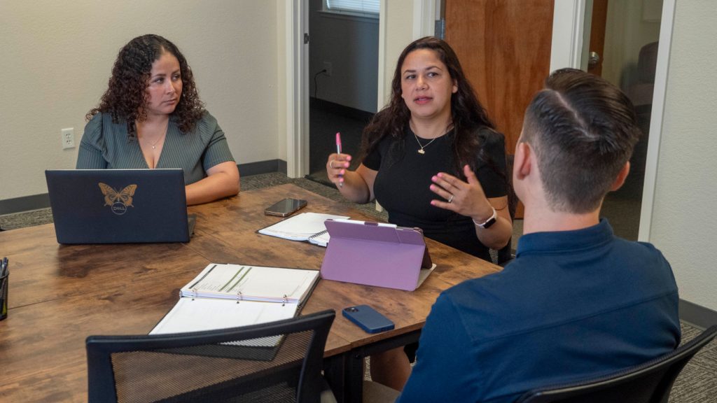 Three people sit around a table during a meeting, engaged in a discussion, with two of them using laptops.