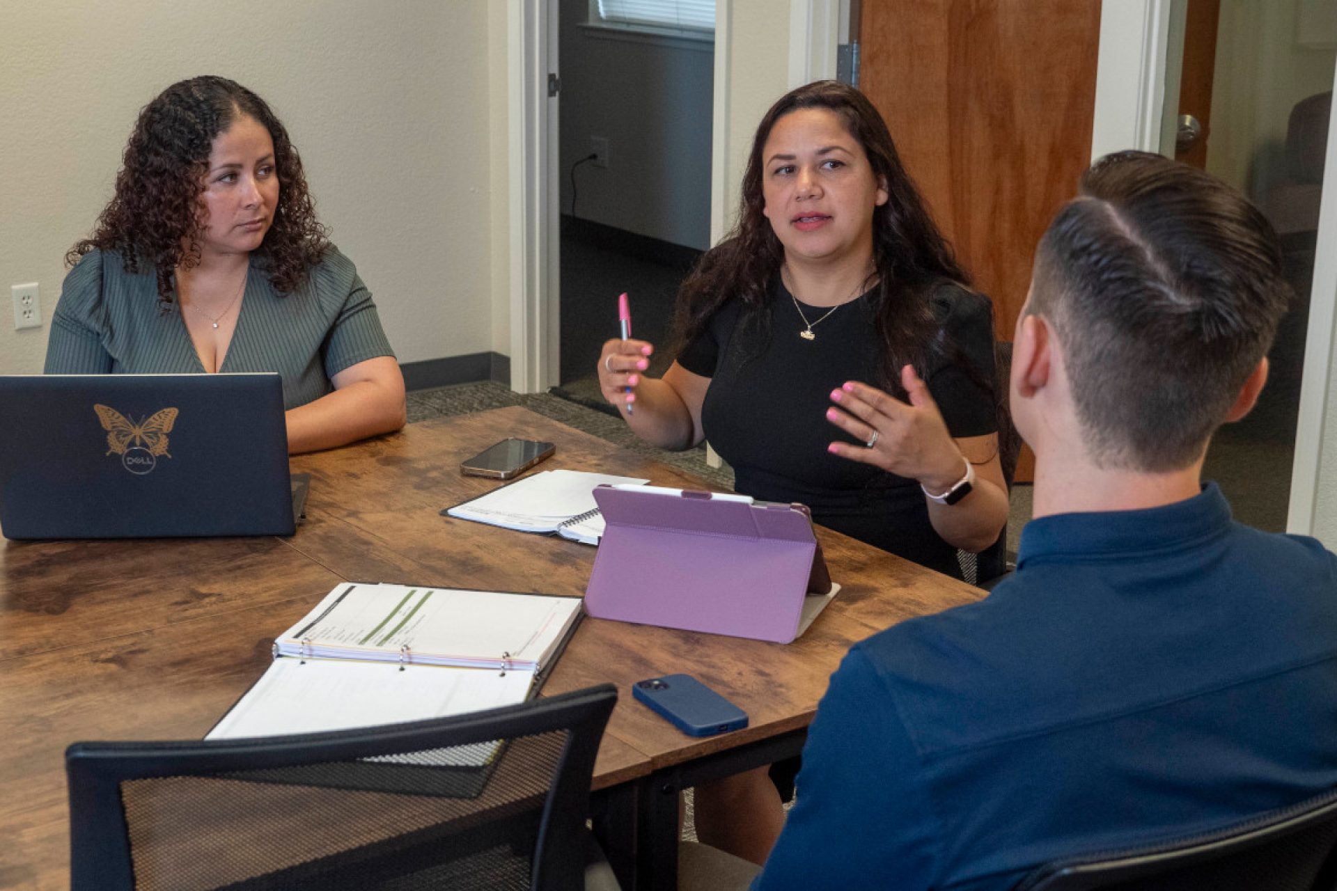 Three people sit around a table during a meeting, engaged in a discussion, with two of them using laptops.