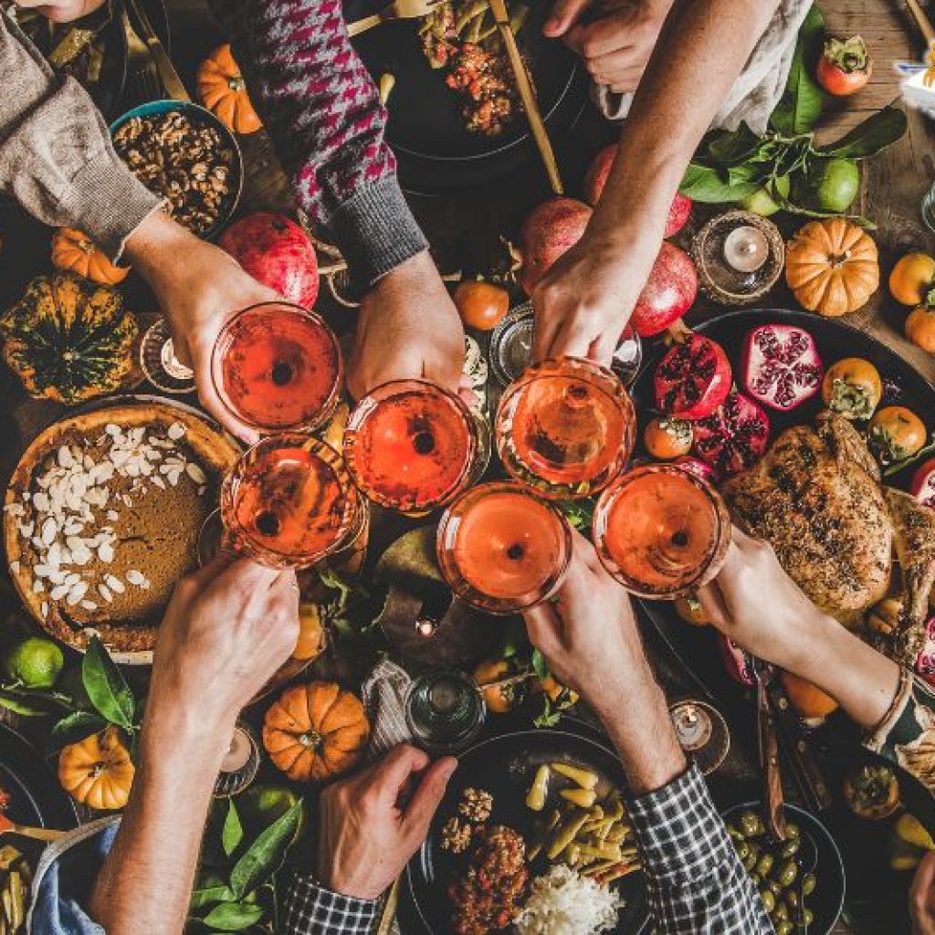 A toast of six glasses filled with an orange beverage help by 6 different people over a large Thanksgiving dinner table.