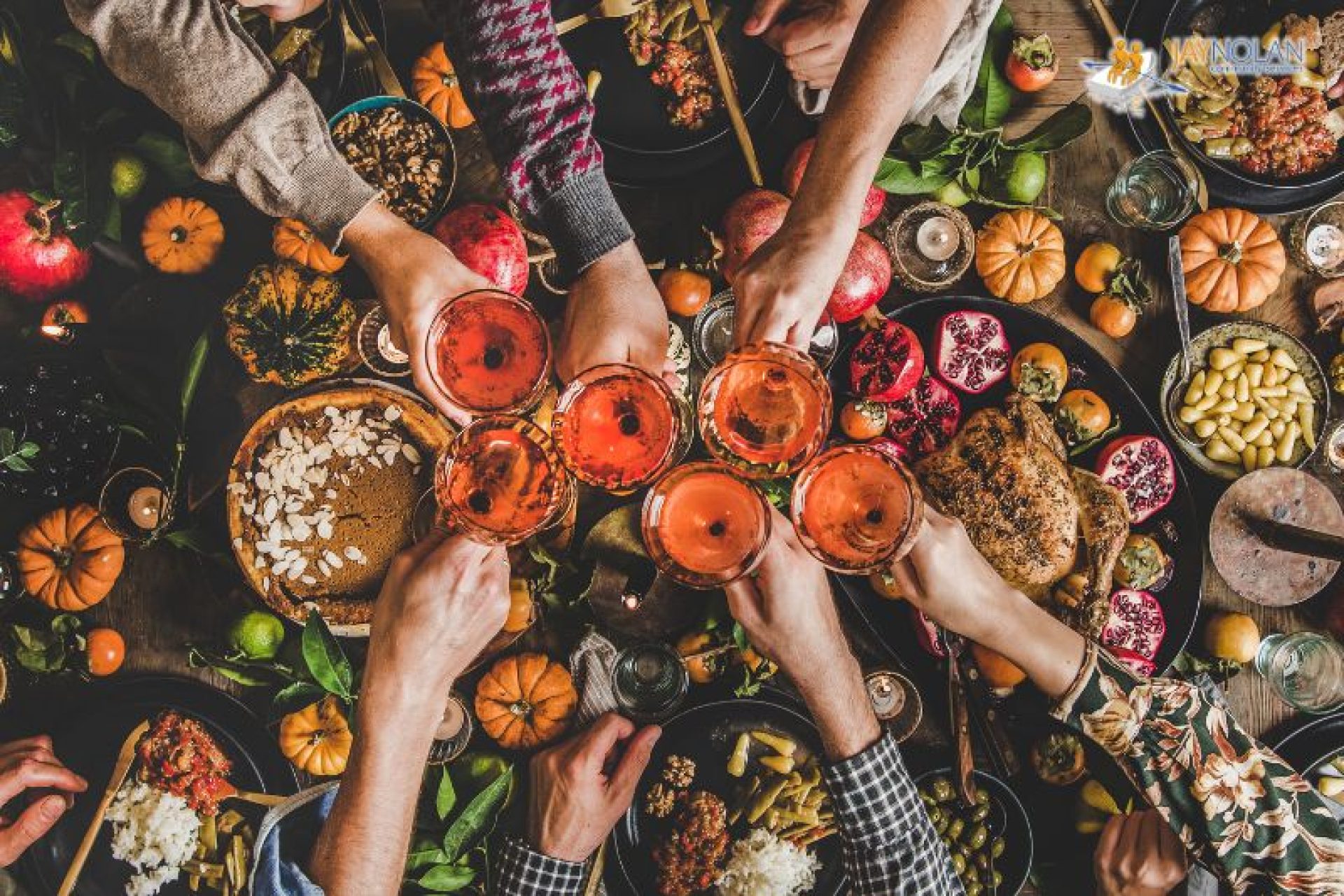 A toast of six glasses filled with an orange beverage help by 6 different people over a large Thanksgiving dinner table.