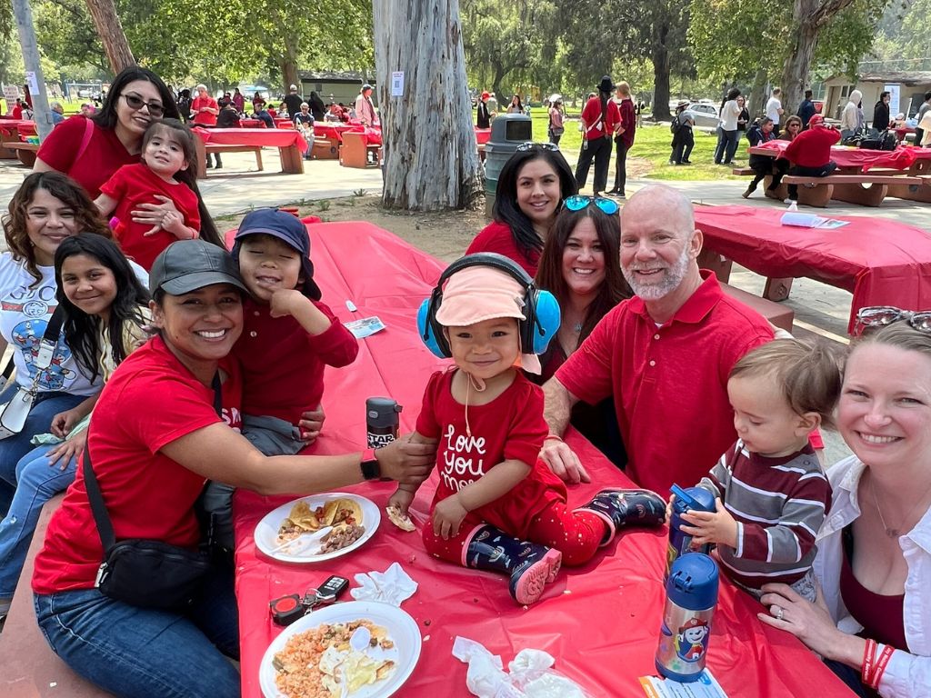 Families with small children all wearing red sitting at a picnic table in the park.