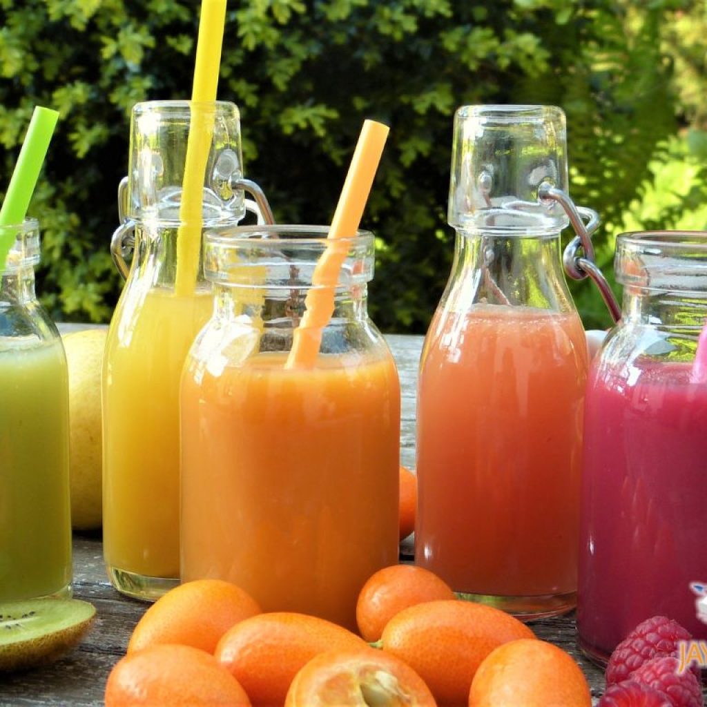 Five glass bottles of different colored juice sitting on a table surrounded by fruit.