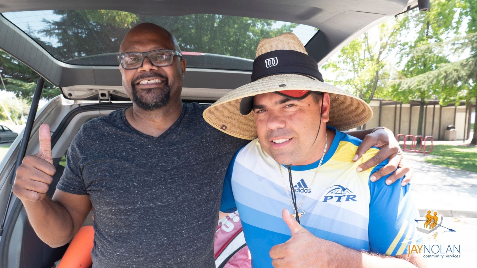 A male African American direct support professional and a Caucasian man who is his client standing by a car while holding their thumbs up.