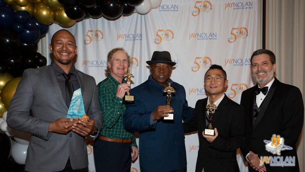 Five male Jay Nolan employees in suits holding awards in front of a balloon arch and a step and repeat banner with the Jay Nolan logo repeated on it.
