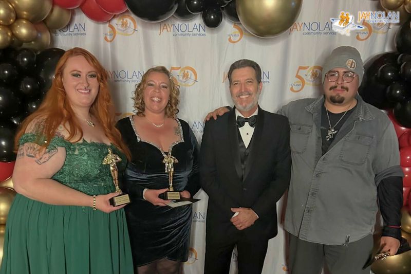 Jay Nolan employees in formal attire holding awards in front of a balloon arch and a step and repeat banner with the Jay Nolan logo repeated on it.