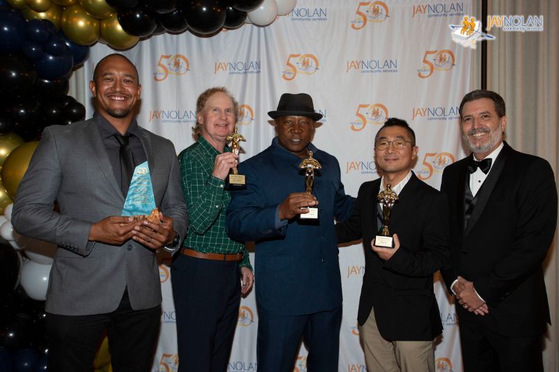 Five male Jay Nolan employees in suits holding awards in front of a balloon arch and a step and repeat banner with the Jay Nolan logo repeated on it.