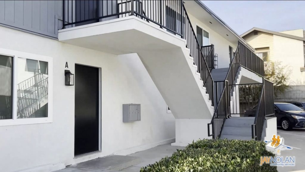Exterior of a two-story white apartment building with stairs on the outside and a hedge out front.