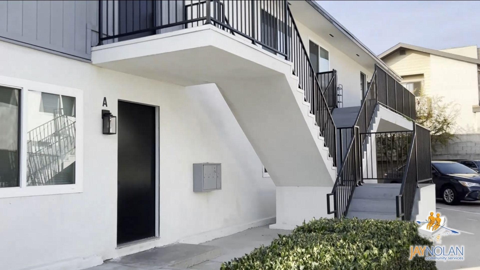 Housing Featured Image Exterior of a two-story white apartment building with stairs on the outside and a hedge out front.