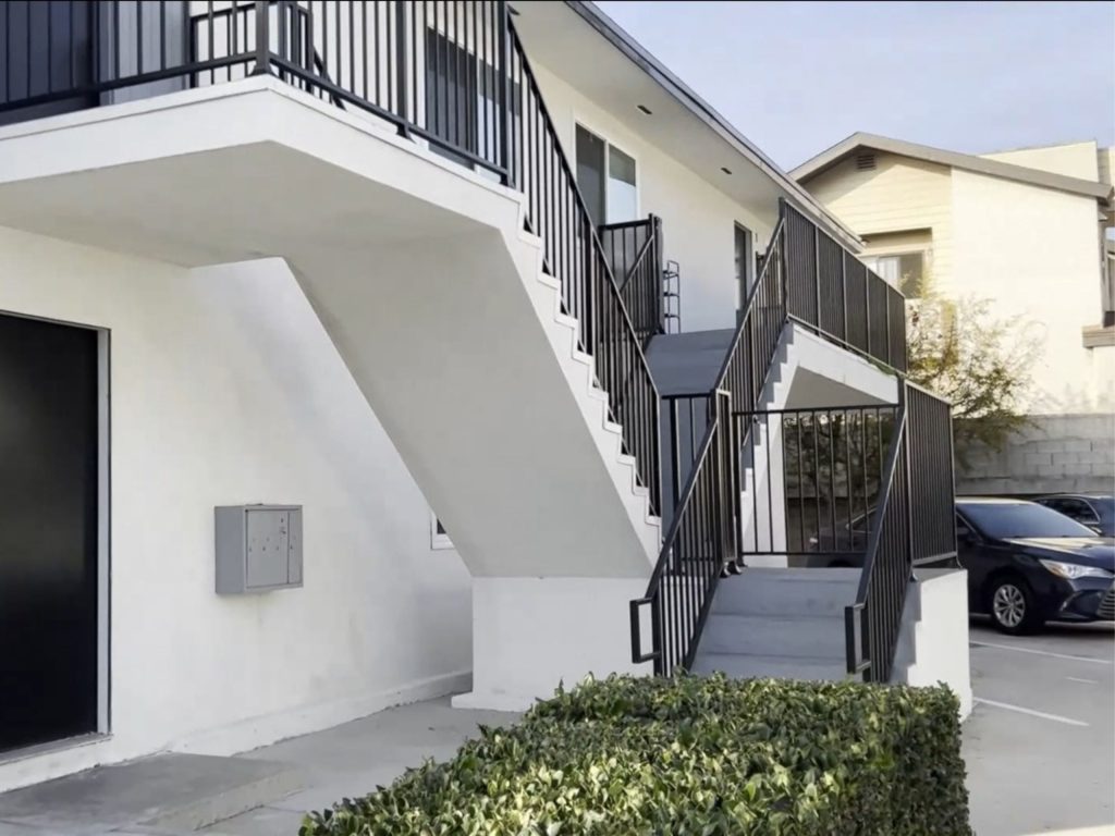 Exterior of a two-story white apartment building with stairs on the outside and a hedge out front.