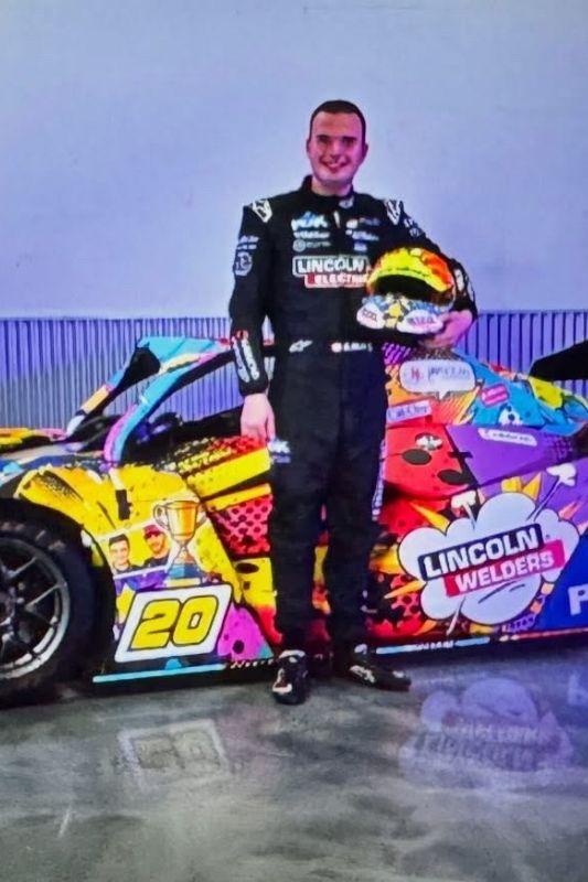 Young man Austin Riley from Racing with Autism standing in front of his 2026 racecar while holding a racecar helmet