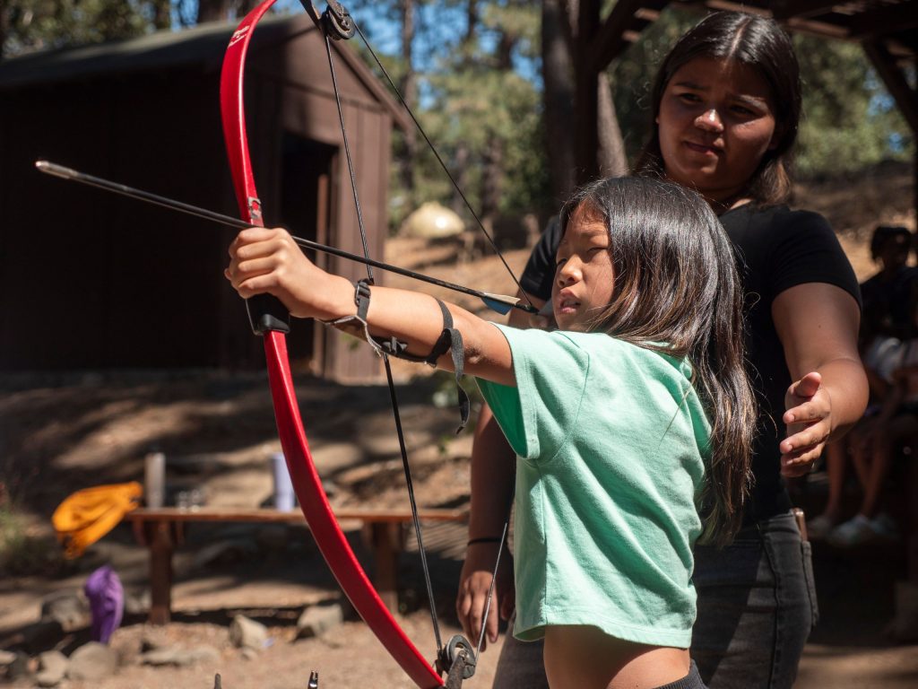 Young girl with a pale green shirt shooting a red bow and arrow at a camp