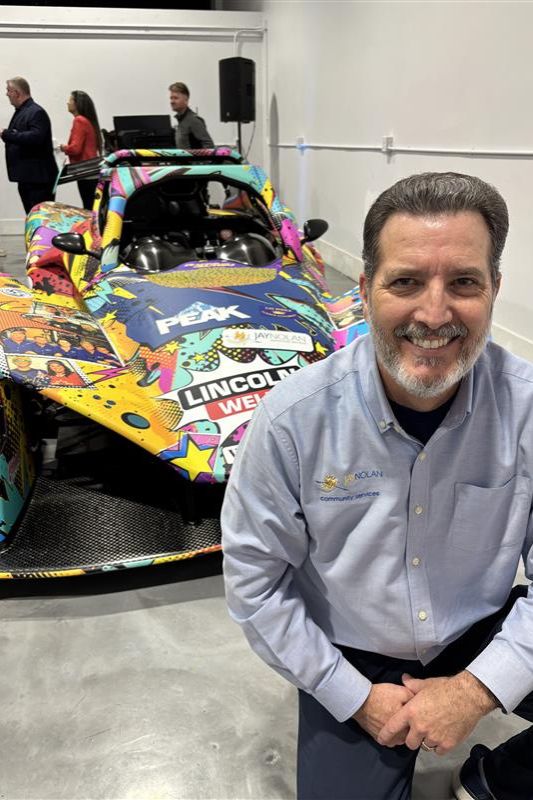 Jay Nolan CEO Edward Amey poses in front of Racing with Autism's 2026 racecar. The Jay Nolan Community Services logo can be seen on the front.