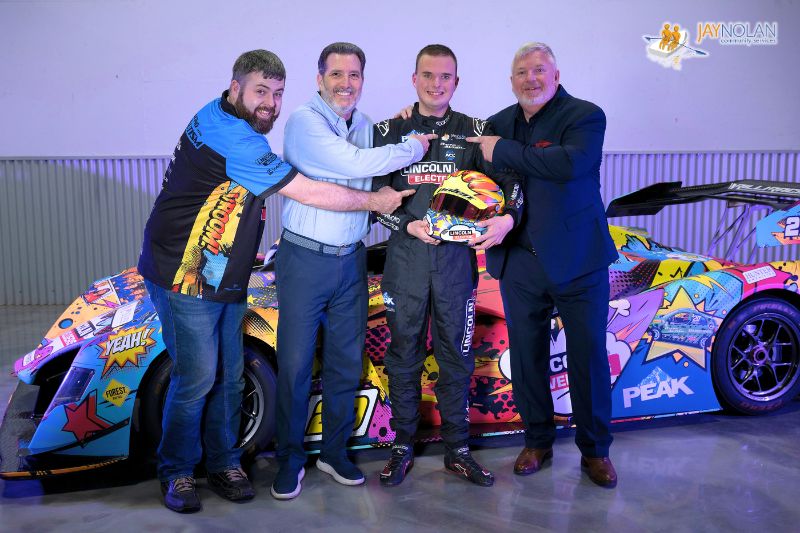 Group photo of racecar driver Austin Riley, Austin's father, CEO of Jay Nolan Edward Amey, and another member of the Racing with Autism team all pointing to the Jay Nolan Community Services logo on Austin's racing uniform.