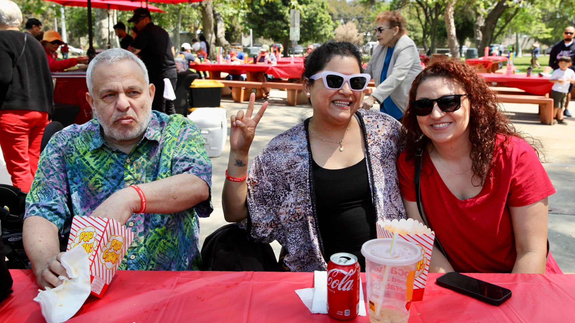Two women wearing sunglasses and a man in a hawaiian shirt sitting at a picnic table with a red tablecloth