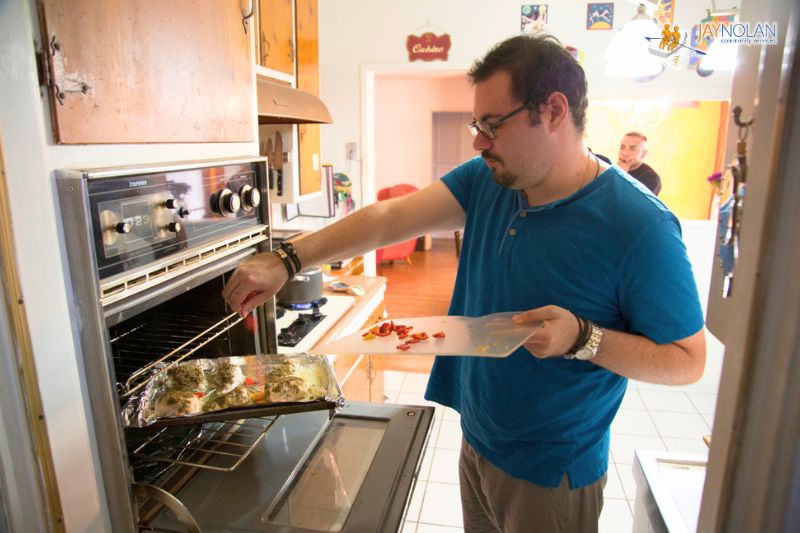Young Caucasian man wearing a blue shirt putting a tray of chicken and vegetables into an oven.
