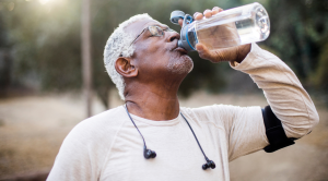 Older adult drinking water from a reusable bottle outdoors while wearing athletic clothing and earbuds highlighting staying hydrated and maintaining healthy habits during warm weather