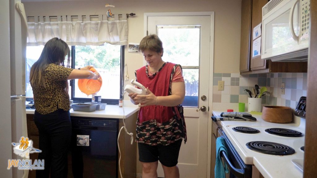 Two women in a kitchen. One of them is holding a hand mixer and the other is washing a bowel in the sink.
