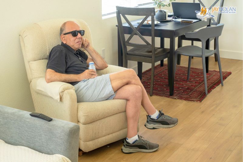 Caucasian man receiving Supported Living Services wearing sungalsses sitting in a white armchair in his living room.