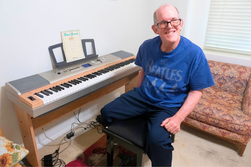 Older Caucasian man receiving Supported Living Services wearing a blue t-shirt and dark pants sitting at a piano in his own home.